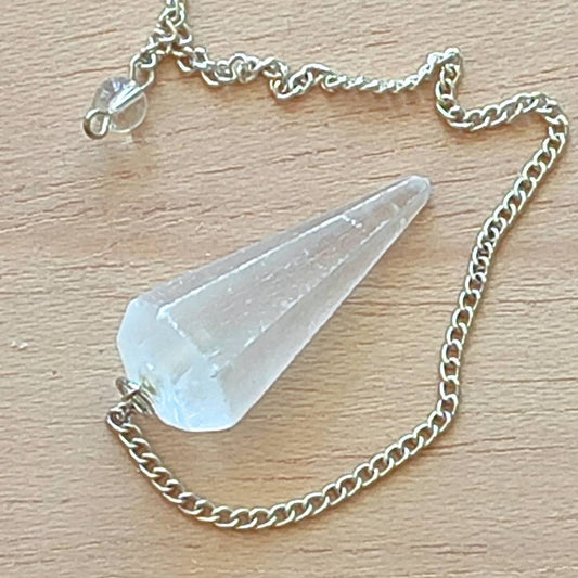 Pêndulo radiestésico em selenita pousado sobre mesa de madeira, representando conexão energética com simplicidade e presença.