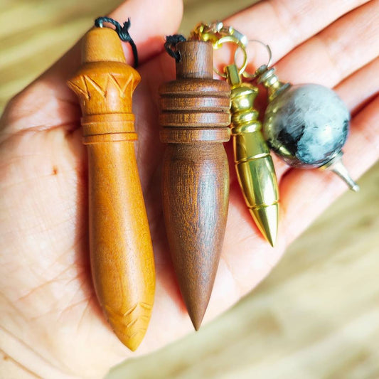Quatro pêndulos radiestésicos exibidos sobre uma mão: dois de madeira (modelos egípcio e frade), um em metal dourado e outro com esfera de cristal natural. Ferramentas vibracionais usadas na prática da RADIESTESIA CONSCIENTE para análise energética.