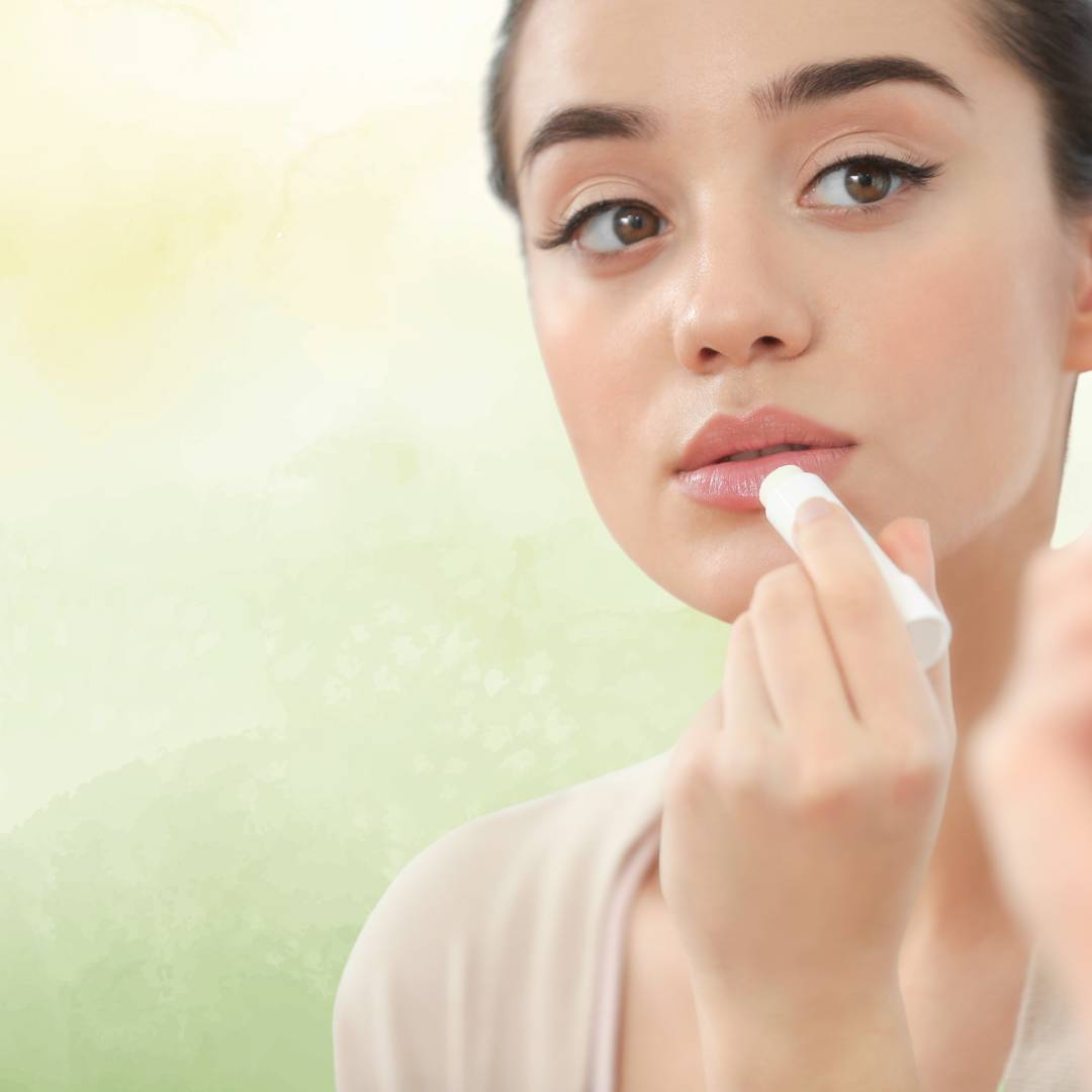 Mulher jovem e radiante, aplicando bálsamo labial em stick, sobre um fundo sereno de cor verde claro, refletindo o cuidado natural da Tilguti.
