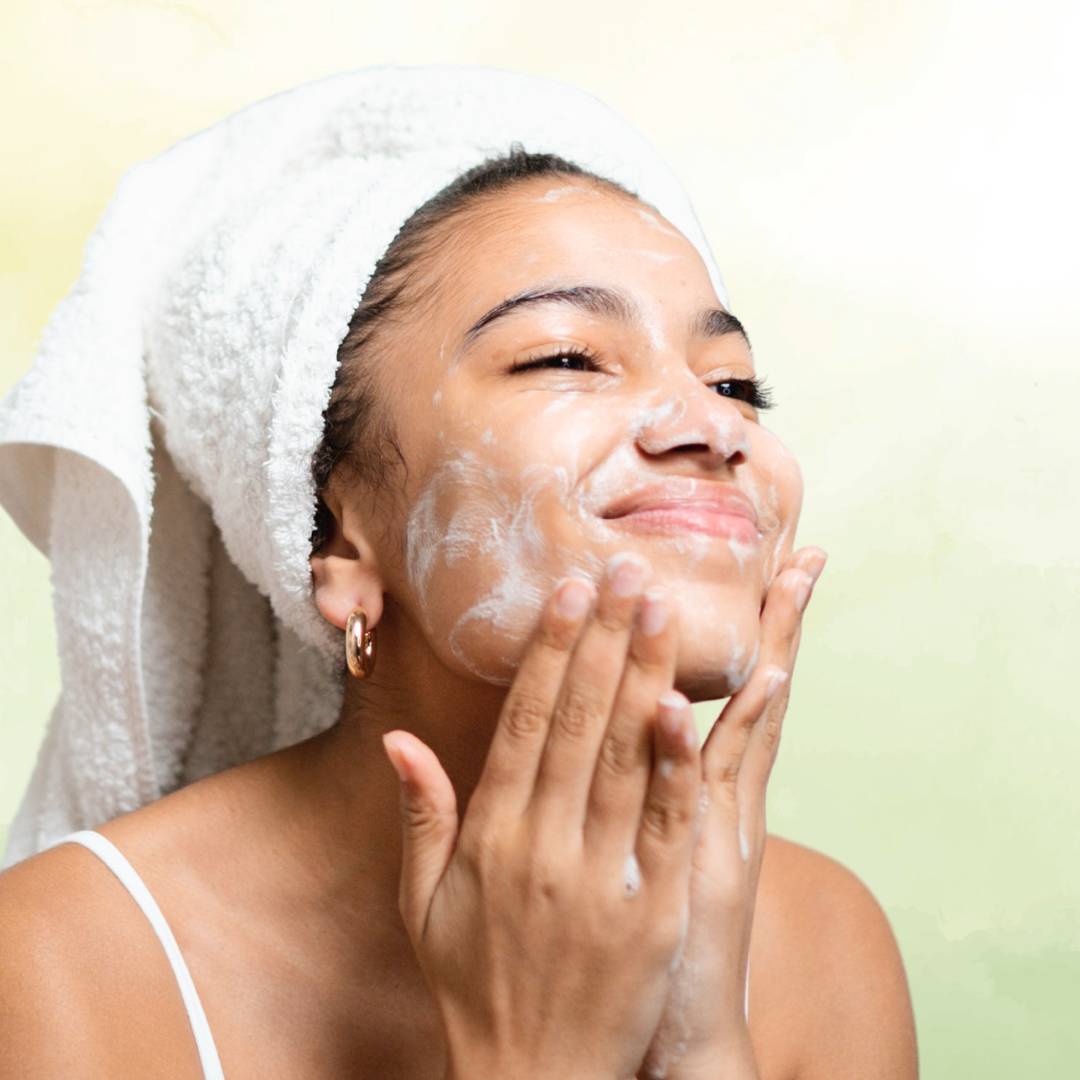 Mulher jovem e radiante, com fundo verde claro, aplicando suavemente leite de limpeza no rosto e cabelo, evidenciando um momento de higiene, cuidado e bem-estar natural.